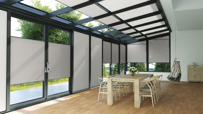 Moderner Wintergarten mit großflächigen Glasflächen und halb heruntergelassenem Wabenplissee-Sonnenschutz, eingerichteter Essbereich mit hellem Holztisch und sechs Stühlen auf hellem Parkettboden.