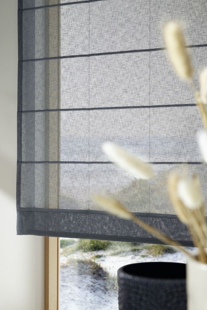 Detailaufnahme eines transparenten Raffrollos in dunklem Grau an einem Fenster mit Blick auf eine verschneite Landschaft, im Vordergrund ein schwarzer Blumentopf mit dekorativem Trockenstrauß.