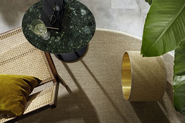 Nahaufnahme einer naturfarbenen Teppichstruktur mit goldenem Lampenschirm und grünem Blatt im Vordergrund