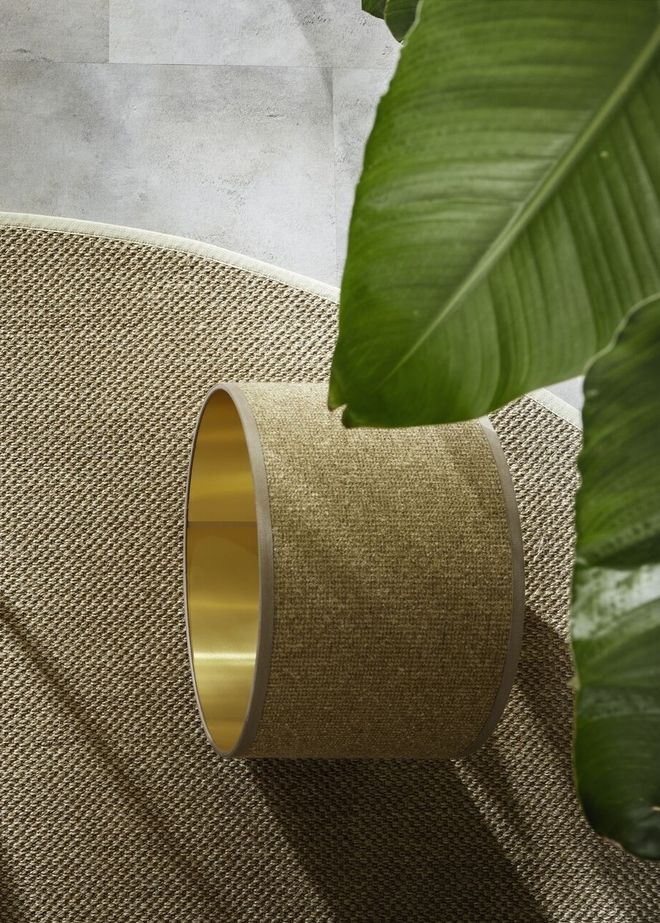 Nahaufnahme einer naturfarbenen Teppichstruktur mit goldenem Lampenschirm und grünem Blatt im Vordergrund