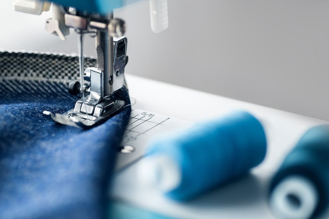 Nahaufnahme einer modernen Nähmaschine beim Nähen von blauem Stoff mit Garnrollen im Vordergrund